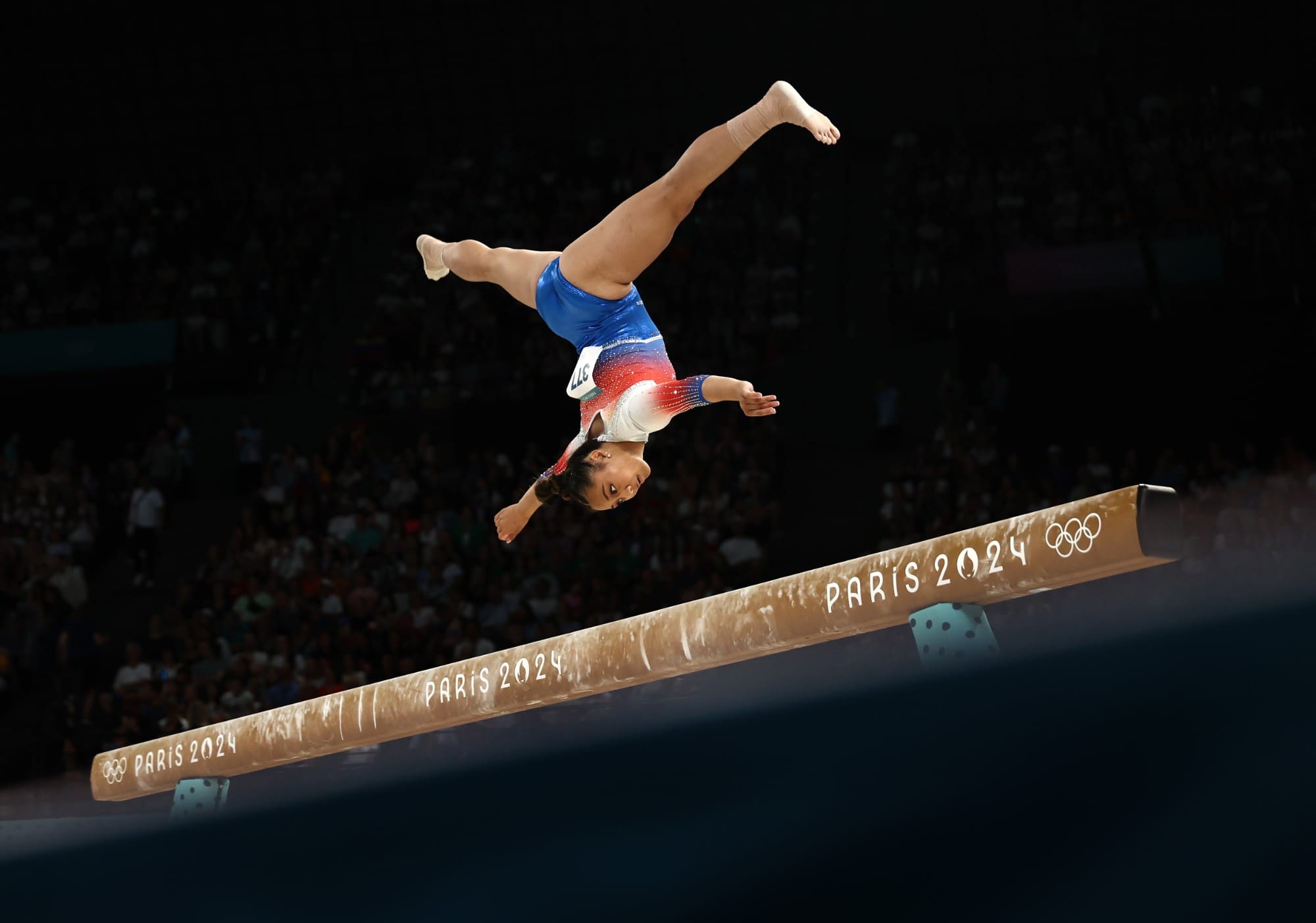 A gymnast upside down in mid-flight above the balance beam at the Paris 2024 Olympic Games.